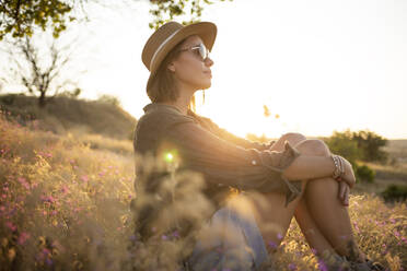 Young woman wearing sunhat, sitting on meadow at sunset - KNTF03085