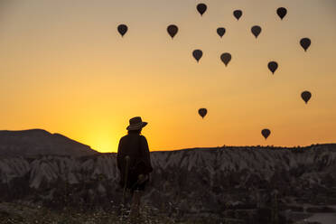 Rear view of silhouette young woman looking at hot air balloons while standing on land in Goreme, Cappadocia, Turkey - KNTF03094