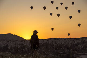 Rückansicht einer jungen Frau mit Silhouette, die Heißluftballons betrachtet, während sie an Land in Goreme, Kappadokien, Türkei steht - KNTF03094