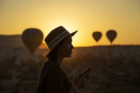 Silhouette einer jungen Frau, die ihr Smartphone benutzt, während sie bei Sonnenuntergang in Kappadokien auf dem Land steht, Türkei, lizenzfreies Stockfoto