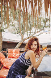 Junge Frau mit einem Getränk sitzt auf einer Bank im Freien und schaut sich um - LJF00767