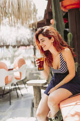 Glückliche junge Frau mit einem Getränk auf einer Bank im Freien sitzend - LJF00771