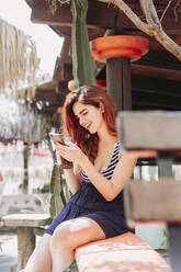 Young woman with a drink and cell phone sitting on a bench outdoors - LJF00772