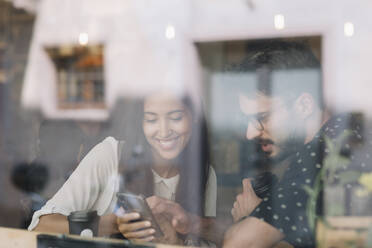 Junges Paar mit Smartphone hinter einer Fensterscheibe in einem Cafe - ALBF00947