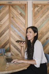 Glückliche junge Geschäftsfrau mit Laptop und Mobiltelefon am Tisch in einem Café - ALBF00968