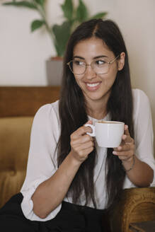 Smiling young woman with cup of coffee sitting in armchair in a cafe - ALBF00977
