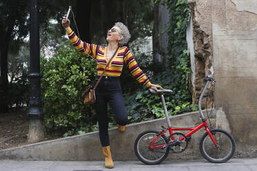 Modische reife Frau mit Fahrrad nimmt Selfie mit Smartphone - RTBF01328
