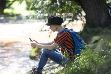 Frau mit Rucksack sitzt am Flussufer und schaut auf ihr Smartphone - SGF02408