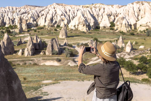 Seitenansicht einer jungen Frau, die mit ihrem Smartphone die felsige Landschaft im Dorf Goreme, Kappadokien, Türkei, fotografiert - KNTF03254