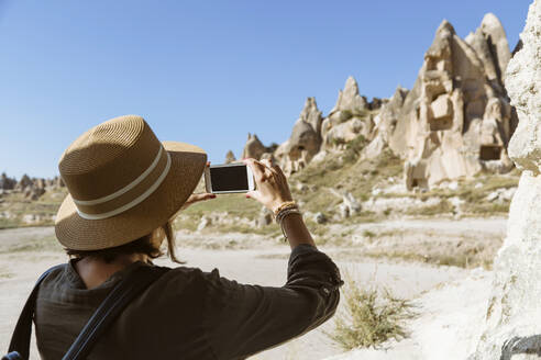 Rückansicht einer jungen Frau, die mit ihrem Smartphone Felsformationen in Goreme, Kappadokien, Türkei, fotografiert - KNTF03264