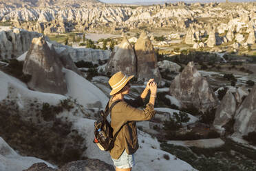 Side view of young woman photographing rock formations with smart phone in Goreme, Cappadocia, Turkey - KNTF03271
