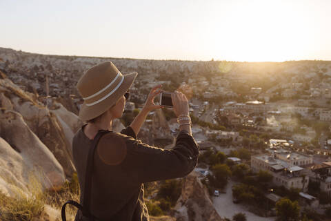 Seitenansicht einer jungen Frau, die während des Sonnenuntergangs mit ihrem Smartphone die Stadt fotografiert, Kappadokien, Türkei, lizenzfreies Stockfoto
