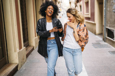 Multikulturelle glückliche Frauen, die in der Stadt spazieren gehen, auf ihr Smartphone schauen und lachen - MPPF00006
