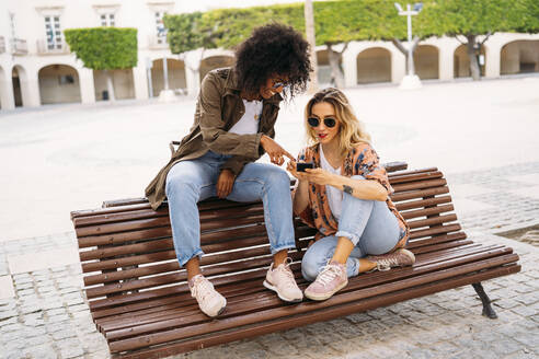 Multicultural happy women talking and using smartphone, sitting on bench - MPPF00013