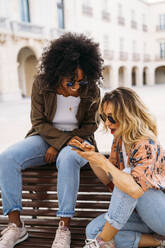 Multikulturelle glückliche Frauen, die sich unterhalten und ein Smartphone benutzen, auf einer Bank sitzend - MPPF00014