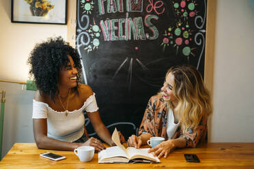 Multicultural women laughing in a cafe, reading a book - MPPF00032