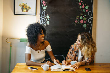 Multicultural women laughing in a cafe, reading a book - MPPF00033