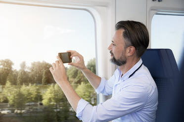 Älterer Mann sitzt in einem Zug und macht Fotos mit seinem Smartphone - FKF03562