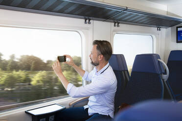 Älterer Mann sitzt in einem Zug und macht Fotos mit seinem Smartphone - FKF03563