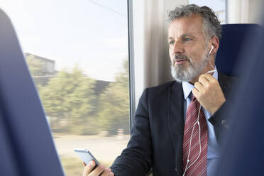 Mature businessman sitting train, using earphones and smartphone - FKF03580