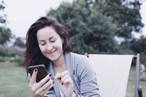 Lächelnde Frau, die auf einem Liegestuhl im Garten ein Mobiltelefon benutzt, lizenzfreies Stockfoto