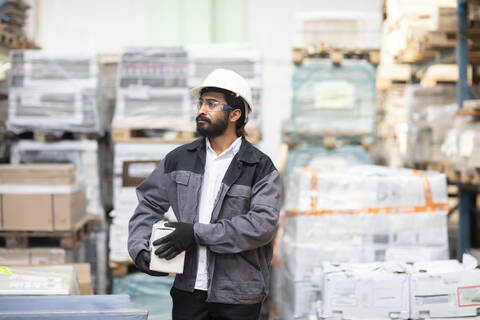 Junger Mann mit Schutzhelm arbeitet in einem Lagerhaus und trägt eine Kiste, lizenzfreies Stockfoto