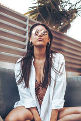 Young woman in shirt and sunglasses sitting on a sofa outside home - ACPF00613