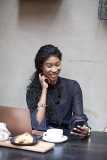 Lächelnde schicke Geschäftsfrau mit Smartphone am Tisch in einem Café - ALBF00985