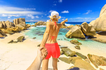 Blonde Touristin im orangefarbenen Bikini hält die Hand ihres Partners an der Anse Marron, Insel La Digue, Seychellen, Indischer Ozean, Afrika - RHPLF06204