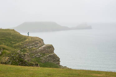 Eine Frau schaut an einem nebligen Tag in Richtung Worms Head auf der Halbinsel Gower, Wales, Vereinigtes Königreich, Europa - RHPLF06291