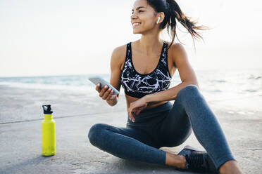 Frau mit Smartphone und In-Ear während des Trainings, sitzend auf einem Steg - OYF00009