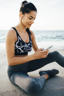 Frau mit Smartphone und In-Ear während des Trainings, sitzend auf einem Steg - OYF00010