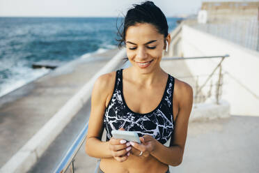 Frau mit Smartphone und In-Ear während des Trainings, sitzend auf einem Steg - OYF00022