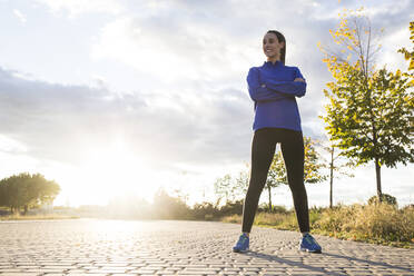 Junge Frau in Sportkleidung, die mit verschränkten Armen im Park posiert und zur Seite schaut - ABZF02551