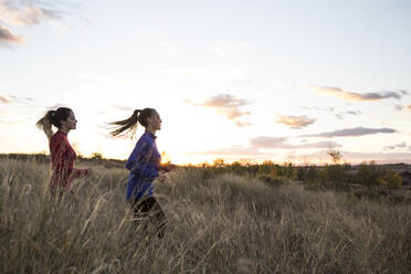 Junge Frauen, die bei Sonnenaufgang gemeinsam über eine Wiese laufen - ABZF02561