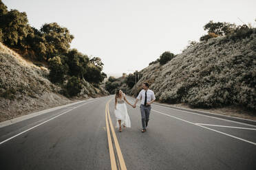 Bride and groom walking hand in hand on a country road - LHPF00752