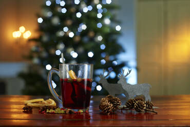 Close-up of mulled wine with food and decorations on wooden table against illuminated Christmas tree at home - KSWF02086