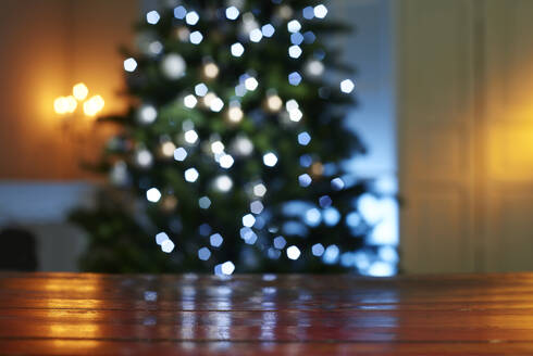 Close-up of wooden table with illuminated Christmas tree in background at home - KSWF02090