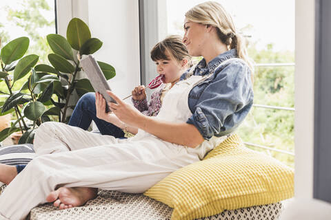 Mutter und Tochter mit Tablet und Lutscher am Fenster zu Hause, lizenzfreies Stockfoto