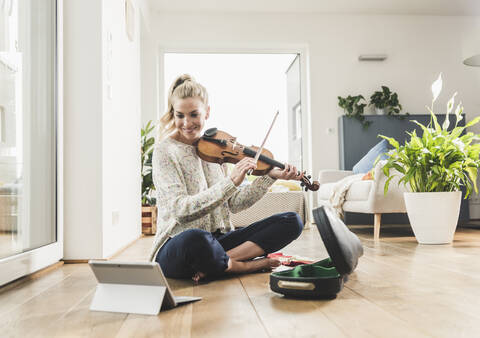 Frau mit Tablet, die zu Hause auf dem Boden sitzt und Geige spielt, lizenzfreies Stockfoto