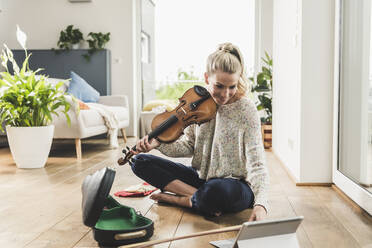 Frau mit Tablet, die zu Hause auf dem Boden sitzt und Geige spielt - UUF18912