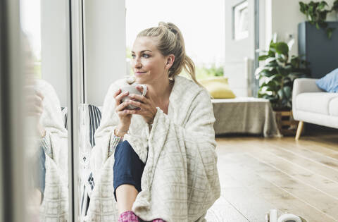 In eine Decke gehüllte Frau, die zu Hause am Fenster sitzt und Kaffee trinkt, lizenzfreies Stockfoto