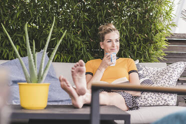 Entspannte Frau sitzt auf der Couch auf der Terrasse zu Hause und trinkt Kaffee - UUF18948