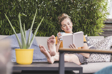 Entspannte Frau sitzt auf der Couch auf der Terrasse zu Hause und liest ein Buch - UUF18949