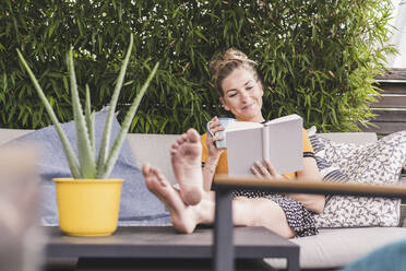 Relaxed woman sitting on couch on terrace at home reading a book - UUF18949