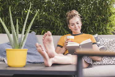 Entspannte Frau sitzt auf der Couch auf der Terrasse zu Hause und liest ein Buch - UUF18950