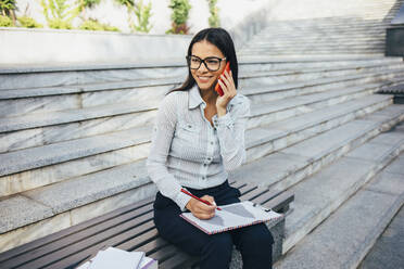 Junge Geschäftsfrau auf einer Bank sitzend, die mit ihrem Handy telefoniert und Notizen macht - OYF00032