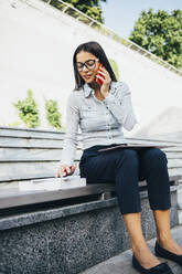 Junge Geschäftsfrau auf einer Bank sitzend, die mit ihrem Handy telefoniert und Notizen macht - OYF00033