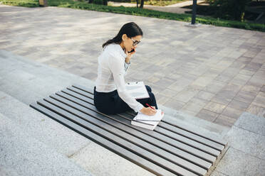 Junge Geschäftsfrau auf einer Bank sitzend, die mit ihrem Handy telefoniert und Notizen macht - OYF00034