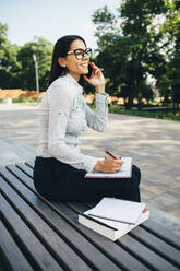 Junge Geschäftsfrau auf einer Bank sitzend, die mit ihrem Handy telefoniert und Notizen macht - OYF00035